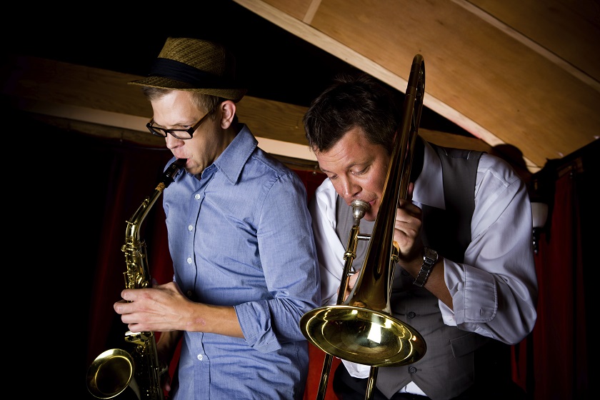 Two hipsters musicians play jazz in a dimly lit restaurant. One has a sax and one has a trombone.
