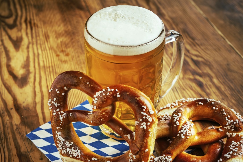 A golden pint of beer sits on a wooden table with two freshly salted pretzels